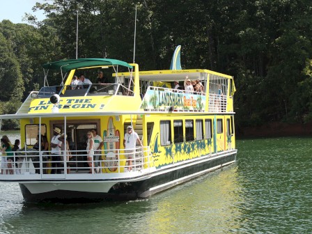 A yellow-and-green boat with people on board, on a lake surrounded by trees, features banners with 