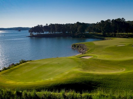 A beautifully manicured golf course beside a calm lake with sunlight reflecting off the water, surrounded by trees, and clear blue sky.