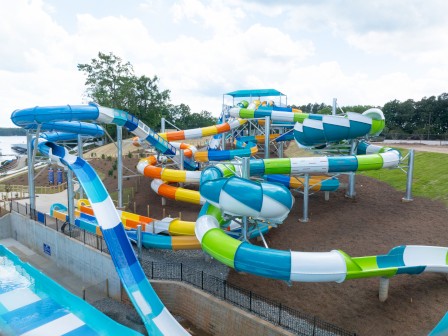 The image shows a colorful water park featuring multiple large water slides with twists and turns, set against a backdrop of trees and a cloudy sky.