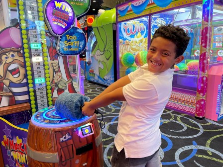 A boy in a white shirt playing a game at an arcade, smiling while interacting with a machine that tests strength or skill, brightly colored machines around.