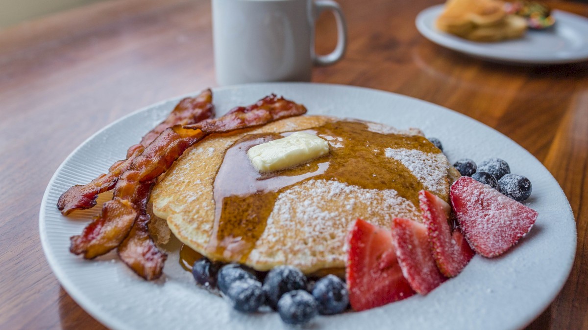 A plate with pancakes, butter, syrup, bacon, strawberries, blueberries, and a sprinkle of powdered sugar, next to a mug of coffee.