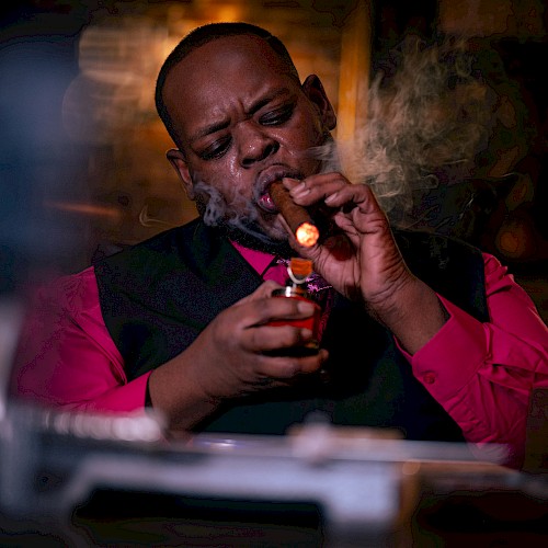 A person in a red shirt lights a cigar in a dimly lit room, with smoke and a relaxed ambiance.