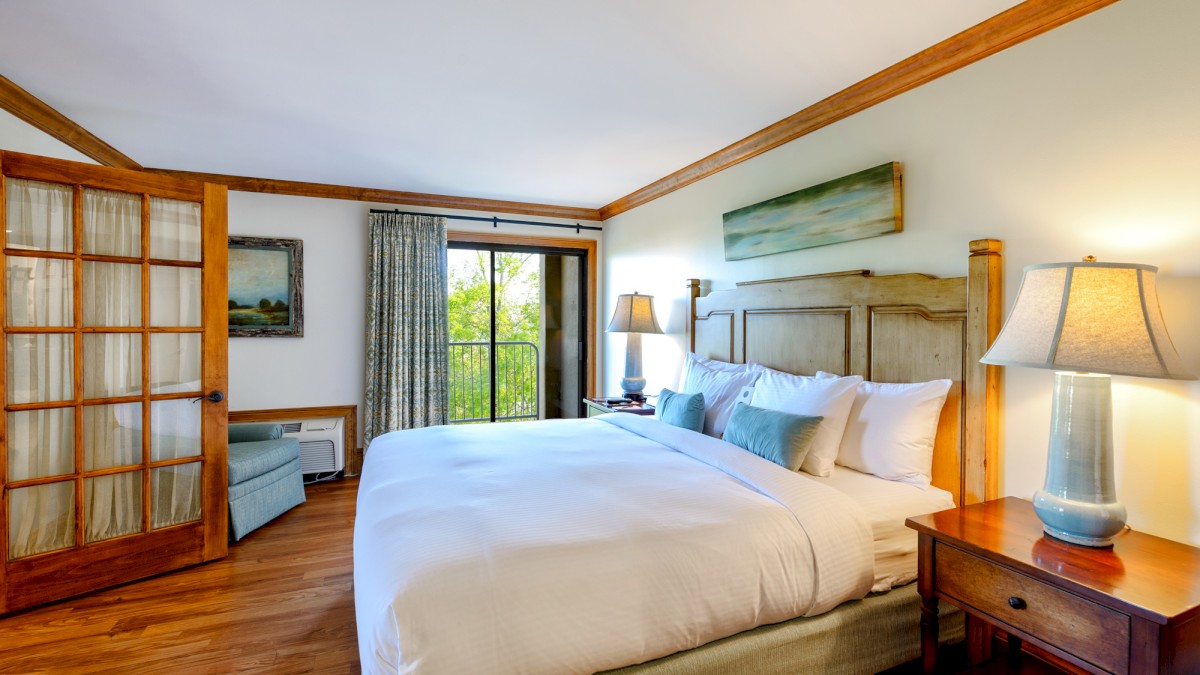 A cozy bedroom features a large bed, wooden furniture, two lamps, artwork, a sliding door, and a window with a scenic view.