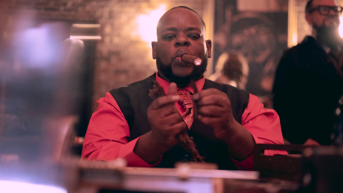 A man in formal attire is cutting a cigar in a warmly lit room with blurry backgrounds.