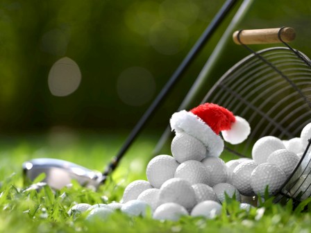 A basket of golf balls with one wearing a Santa hat lies on grass beside a golf club, creating a festive golfing scene.