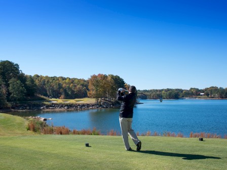 A golfer swings on a lush green course by a calm blue lake, trees in autumn colors lining the water under a clear sky.
