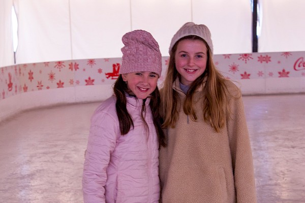 Two girls pose together on an ice skating rink, each wearing winter clothes and ice skates, smiling for the camera.