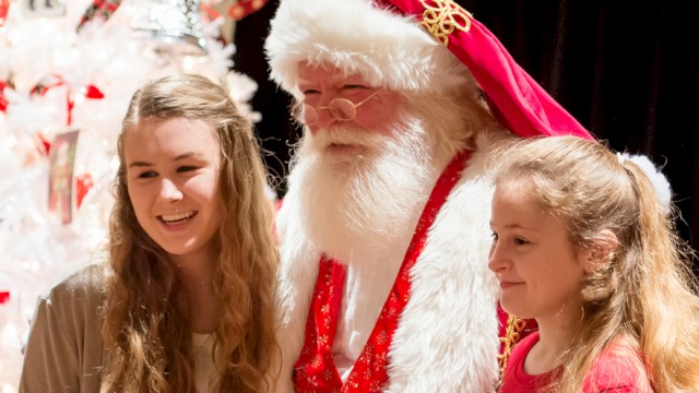 Two young girls are posing with a man dressed as Santa Claus in front of a festive Christmas tree, all smiling happily.