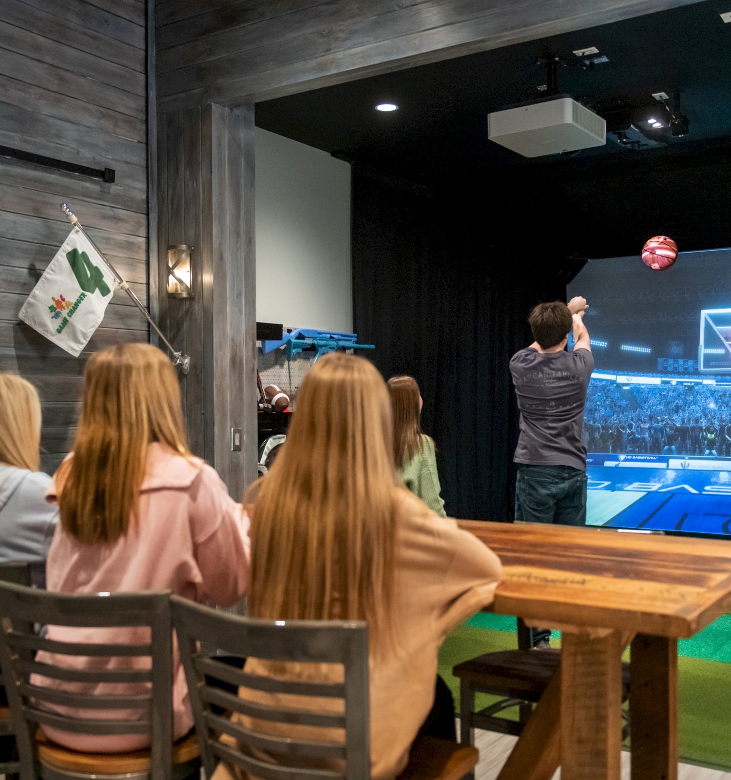 A man is playing a basketball simulation game on a large screen while a group of people watches from behind a wooden table.