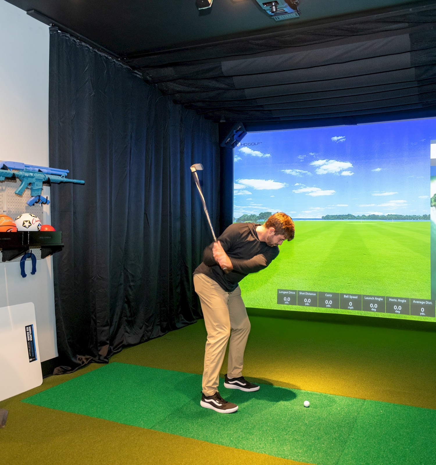 A person is playing golf in an indoor simulator room with equipment on shelves and a virtual course displayed on a screen.