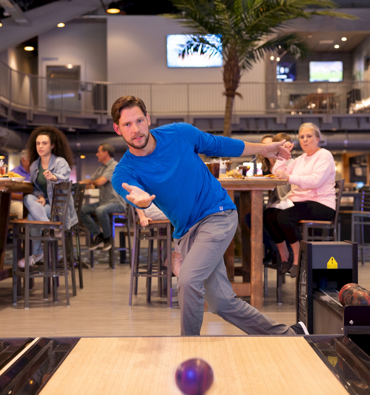A man wearing a blue shirt is bowling, with people watching nearby in a modern alley with tables.