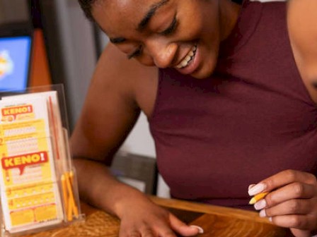 Two people sitting at a table, with one holding a pencil, possibly playing Keno. A drink and a Keno pamphlet with pencils are on the table.
