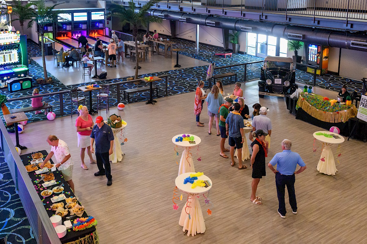 A group of people is attending a social event in a large room with games, decorations, and food tables.