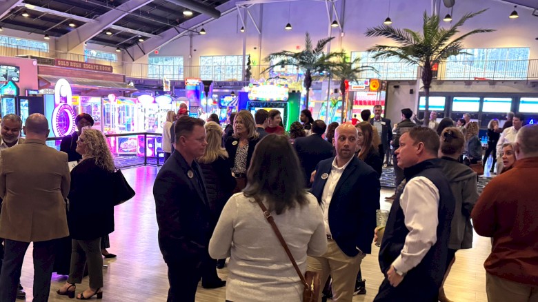 People mingle at an indoor event or expo, chatting in small groups amid colorful booths and decorative palm trees.