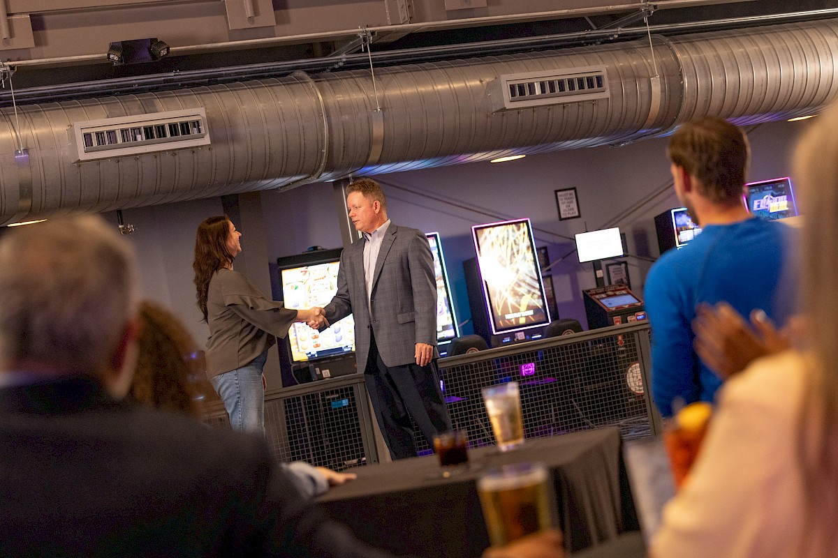 A man and woman shake hands in a modern office or arcade setting, while others sit and watch, enjoying drinks and a casual celebration.