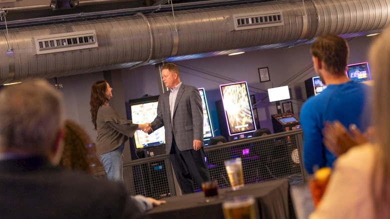 A man and woman shake hands in a modern office or arcade setting, while others sit and watch, enjoying drinks and a casual celebration.