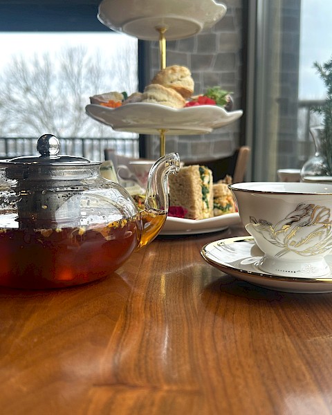A table with a teapot, teacup, and a tiered tray with scones and sandwiches, set in a cozy indoor setting by a window.
