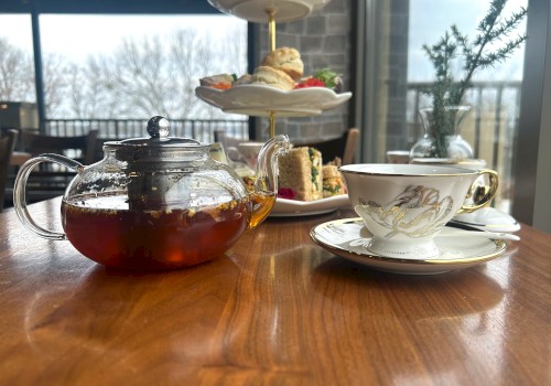 A table with a teapot, teacup, and a tiered tray with scones and sandwiches, set in a cozy indoor setting by a window.