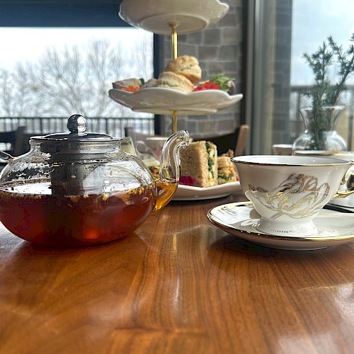 A table with a teapot, teacup, and a tiered tray with scones and sandwiches, set in a cozy indoor setting by a window.