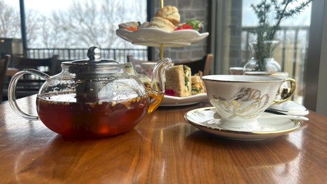 A table with a teapot, teacup, and a tiered tray with scones and sandwiches, set in a cozy indoor setting by a window.