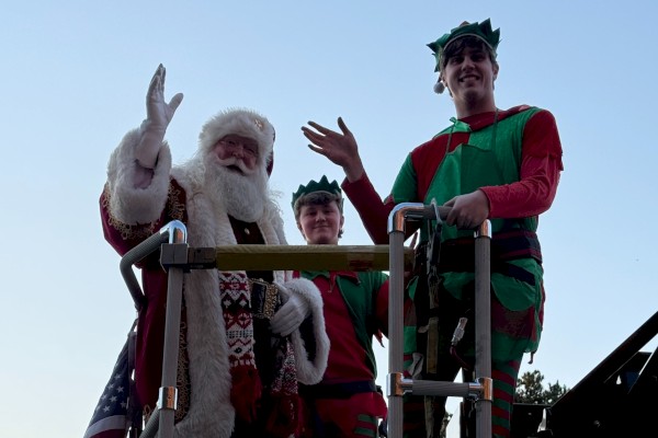 A person in a Santa suit and two others dressed as elves are standing on a fire truck platform, waving.