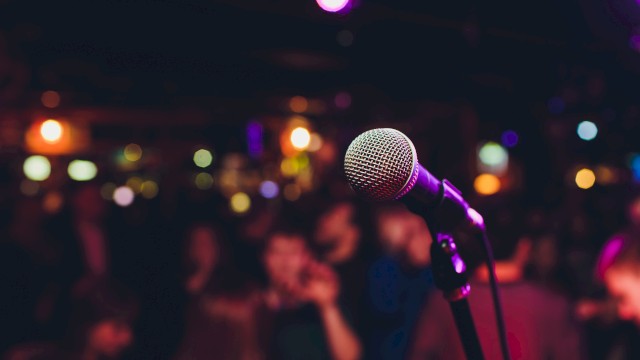 A microphone is in focus against a blurred backdrop of a crowd and colorful lights, suggesting a lively performance or event.