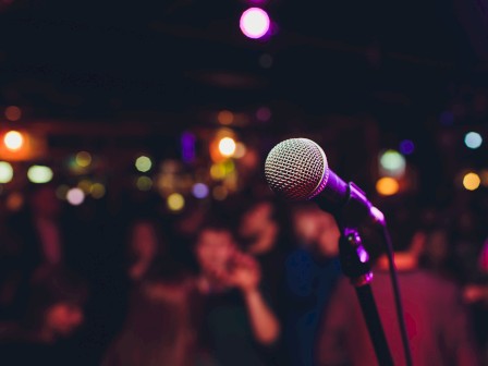 A microphone is in focus on a stand, with a blurred background of colorful lights and people, suggesting a lively event or performance.