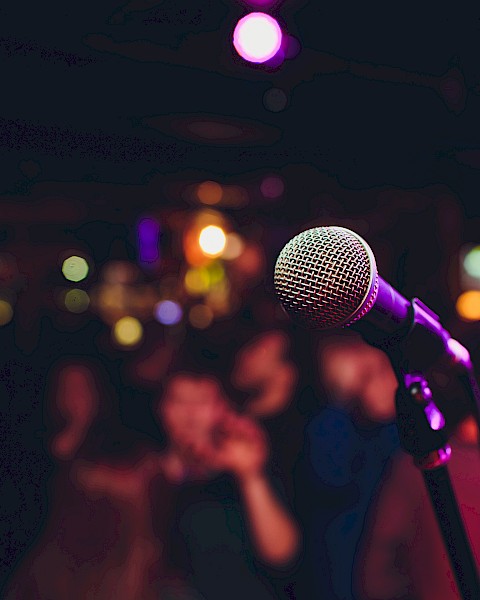 A microphone stands in focus on stage, with a blurred background of colorful lights and a crowd, suggesting a live performance setting.
