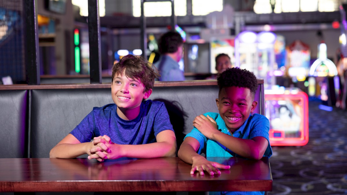 Two kids are sitting at a table in an arcade, smiling and posing for the camera with games in the background.