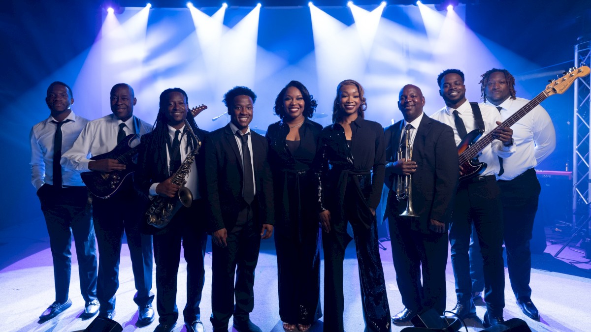 A lively, coordinated band on stage in formal attire with instruments (guitars, sax, bass) under bright stage lights, posing for a group photo.