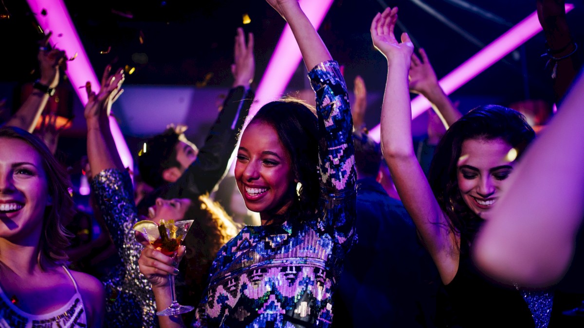 People dancing at a colorful nightclub, raising hands, neon lights, and drinks in hand, celebrating the night.