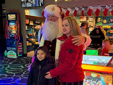 A person dressed as Santa Claus poses with a woman and a child in a colorful arcade, under a sign that reads 