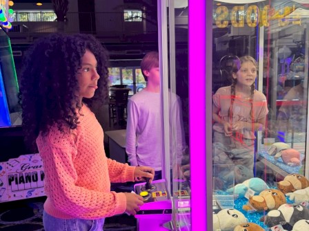 A person is using a claw machine filled with stuffed animals in an arcade, surrounded by colorful lights.