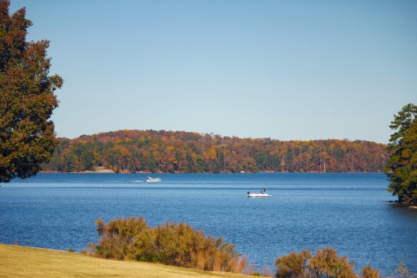 A serene lake scene with two boats on the water, surrounded by trees with autumn foliage under a clear blue sky.