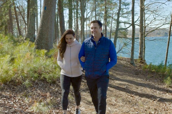 A couple is walking together on a wooded path by a lake, surrounded by trees and sunshine.