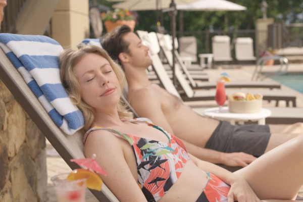 A woman rests on a lounge chair with her eyes closed, while a man relaxes beside her by the pool on a sunny day.
