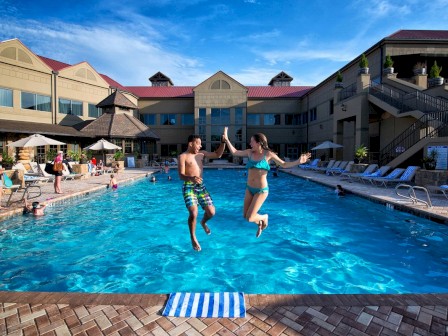 Two people are mid-air, jumping into a swimming pool. There are lounge chairs with umbrellas around the pool and a large building in the background.