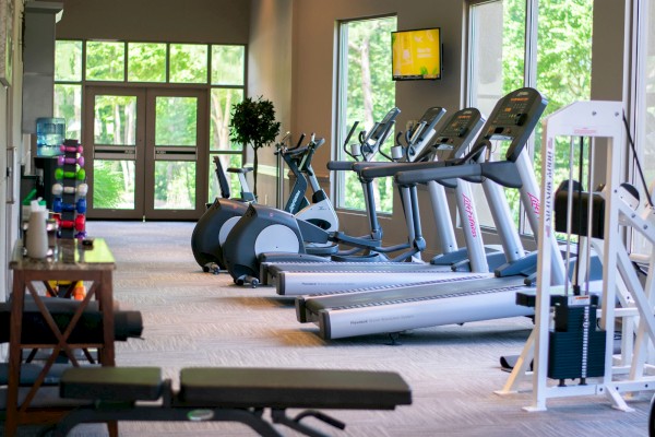The image shows a gym with treadmills, exercise bikes, a weight machine, and benches, with windows and a TV on the wall.