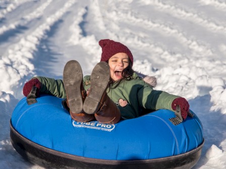A girl in winter clothing smiles and slides down on a snow tube in snowy landscape.