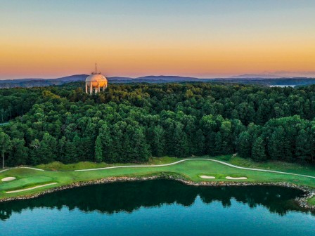 A golf course by a lake at sunset with a domed building and wooded area in the background, under a colorful sky.