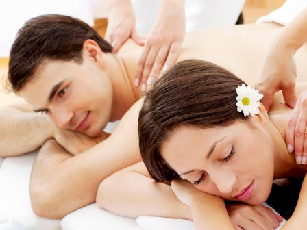 Two people are lying side by side receiving massages. One has a flower in their hair and they both appear relaxed with masseuses working on them.