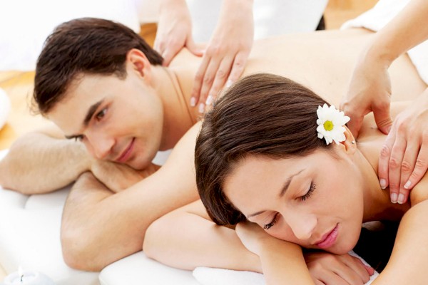 Two people are lying side by side receiving massages. One has a flower in their hair and they both appear relaxed with masseuses working on them.