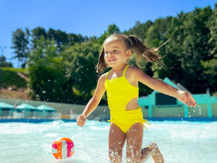 A young girl in a yellow swimsuit joyfully plays in a swimming pool on a sunny day.