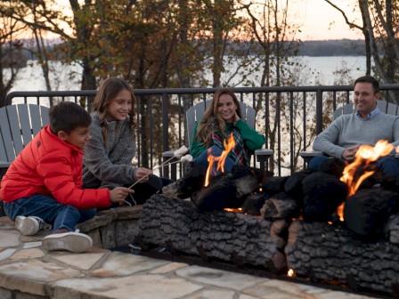 A family of four, including children, are sitting by a fire outdoors in autumn, roasting marshmallows and enjoying a cozy evening.