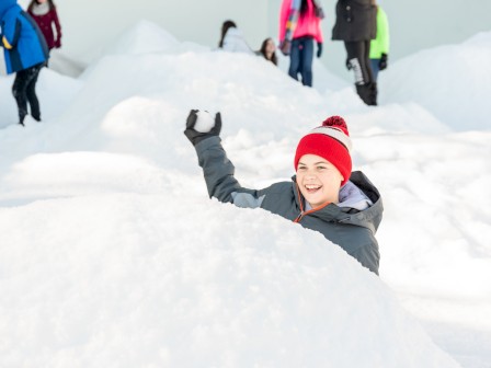 A boy in a red hat and gray jacket enjoys playing in the snow while others gather in the background.