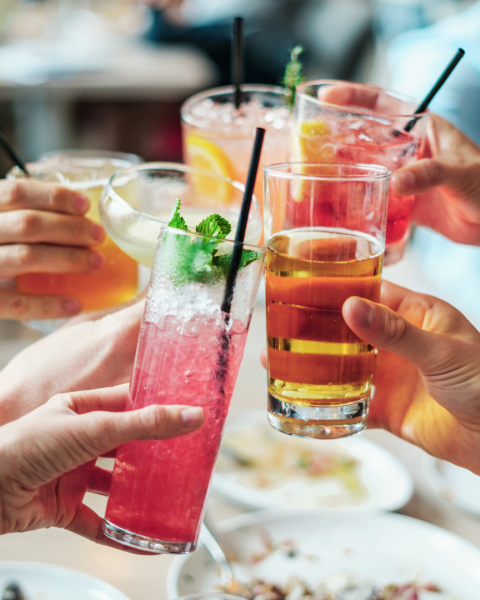 A group of people toasting with assorted colorful drinks, garnished with fruits and herbs, using various glasses and straws.