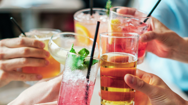 A group of people toasting with assorted colorful drinks, garnished with fruits and herbs, using various glasses and straws.