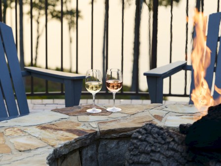 Two wine glasses resting on a stone patio beside a lit fire pit, with blue Adirondack chairs and a railing in the background.