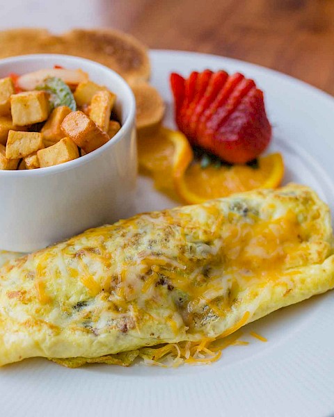 The image shows an omelet on a plate, served with a bowl of diced potatoes, sliced strawberries, orange slices, and pancakes.