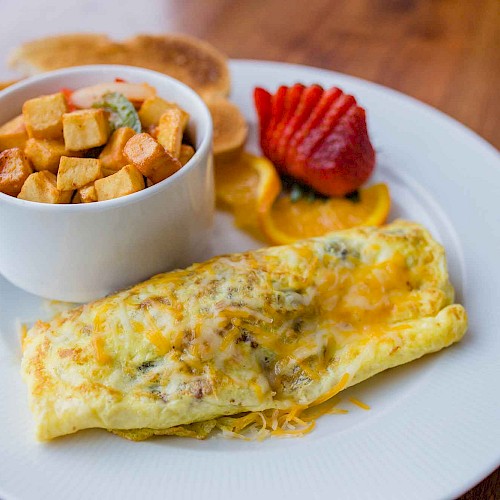 The image shows an omelet on a plate, served with a bowl of diced potatoes, sliced strawberries, orange slices, and pancakes.